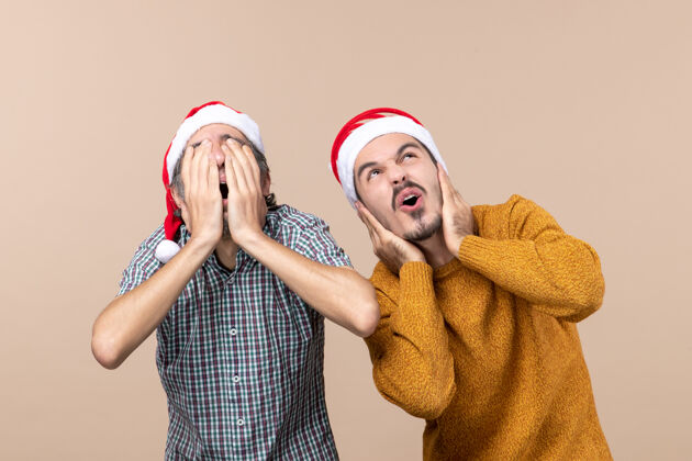 肖像正面图两个戴着圣诞帽的吓坏了的家伙 一个蒙着脸 另一个耳朵在米色的孤立背景上掩护米色帽子