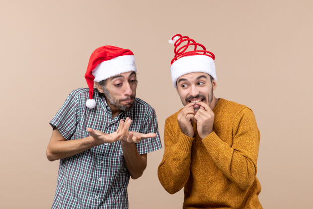 男性正面图两个戴着圣诞帽的好奇的家伙在米色孤立的背景下闲聊伙计们两个帽子