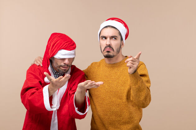 脸前视图两个年轻人一个在看某处 另一个在米色孤立的背景下用圣诞帽遮住眼睛男人肖像男人