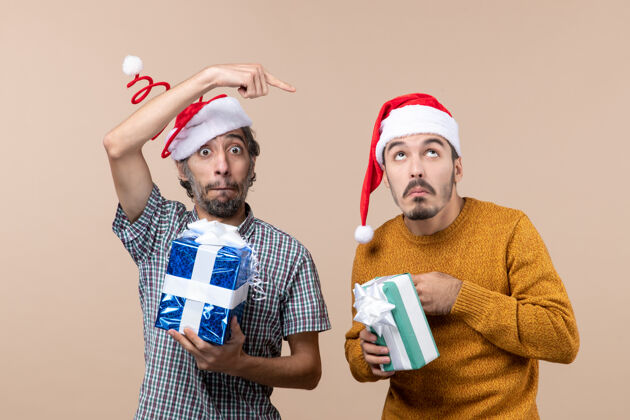 男人前视图两个困惑的家伙拿着圣诞礼物一个手指点显示在米色孤立的背景上的其他米色手指困惑