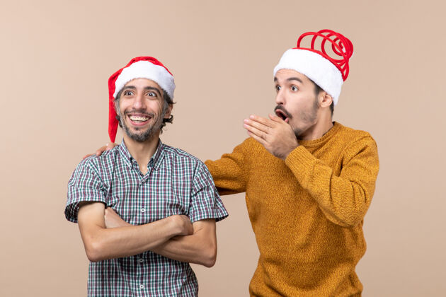 肖像前视图：两个圣诞老人戴着圣诞帽 一个在米色背景上向另一个展示东西帽子圣诞老人视图