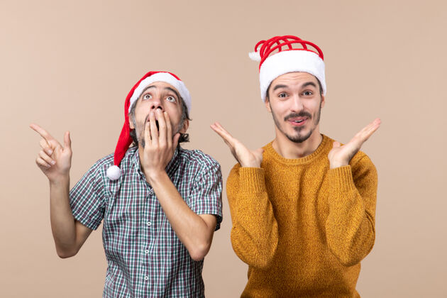 手正面图两个戴着圣诞帽的惊讶的家伙一个看着高处另一个张开双手站在米色的孤立背景上看两个惊奇的家伙两个