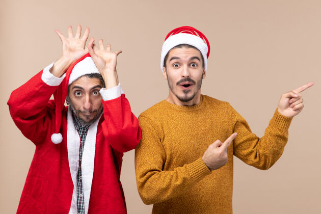 视图正面图两个戴着圣诞帽的朋友 一个举起手来举着头 另一个在米色孤立的背景上显示方向前面肖像男性