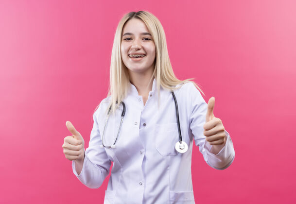 牙科微笑的医生年轻的金发女孩穿着医用长袍 戴着听诊器和牙科支架 在孤立的粉红色背景上竖起大拇指微笑年轻听诊器