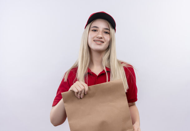 女孩带着微笑的小女孩穿着红色t恤 戴着帽子 戴着牙套 在孤立的白色背景上拿着纸袋口袋纸戴着