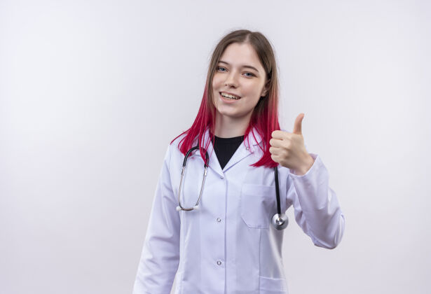 女孩微笑的年轻女医生穿着听诊器医用长袍 在孤立的白色背景上竖起大拇指穿着向上听诊器