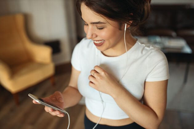 互联网高角度观看可爱欢快的少女穿着白色紧身上衣与朋友聊天或听新的音乐曲目使用在线应用程序在手机上家庭移动耳机