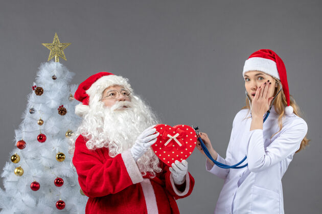 礼物圣诞老人和女医生在灰色墙上用听诊器观察的正视图圣诞十二月医生