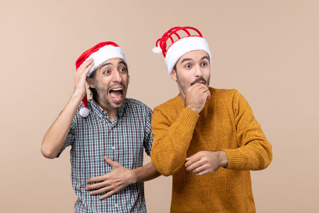 视图正面图两个戴着圣诞帽的惊讶的家伙 一个手放在头上 另一个脸贴在米色孤立的背景上手米色头