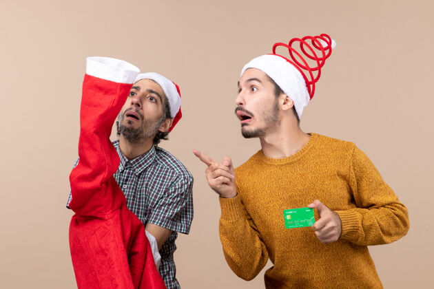 米色前视图两个家伙一个拿着圣诞老人的外套 另一个拿着信用卡看着米色背景上的东西帽子男人拿着