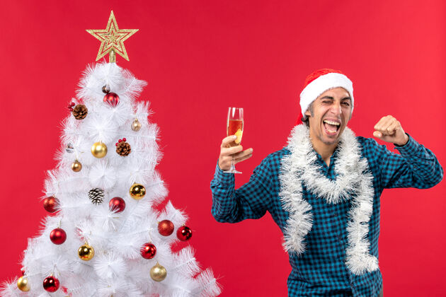 新年前夜圣诞节心情与情绪骄傲疯狂的年轻人与圣诞树附近拿着一杯葡萄酒的蓝色条纹衬衫圣诞老人帽子聚会年轻人年