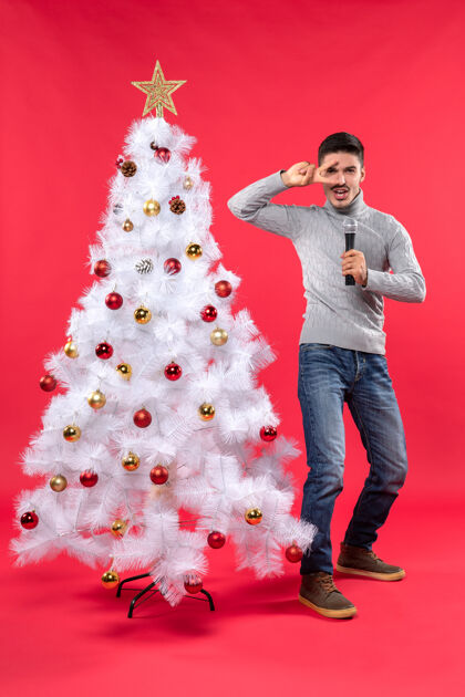 庆祝圣诞节的气氛与自信的家伙穿着牛仔裤站在装饰圣诞树附近 手持麦克风 并显示有趣的手势快乐装饰男人