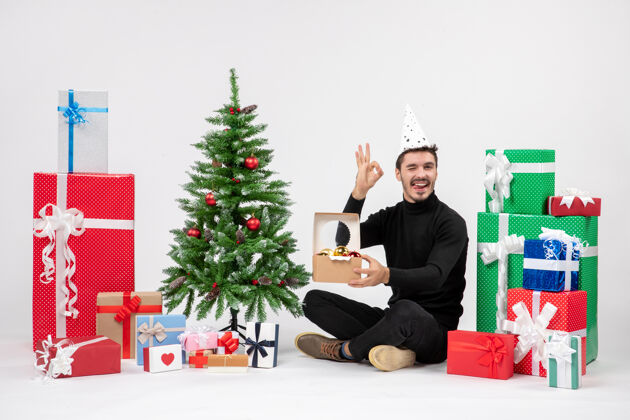 节日正面图：年轻人围坐在节日礼物旁边 手里拿着白色墙上的玩具包快乐人圣诞树
