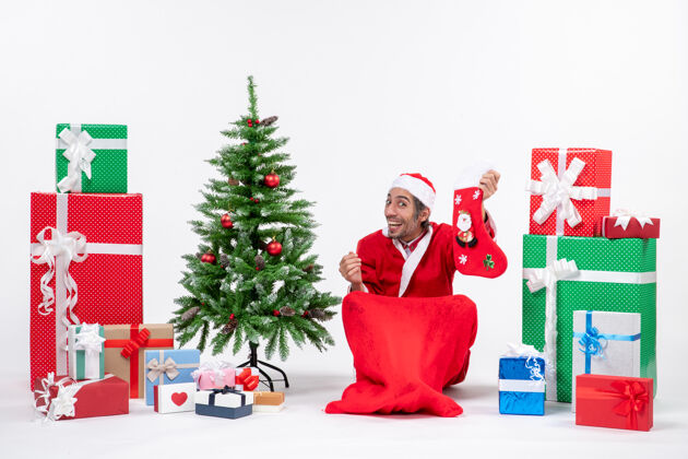 圣诞老人惊喜积极兴奋的圣诞老人坐在地上 拿着圣诞袜靠近礼物和白色背景装饰的新年树圣诞老人拿着惊讶