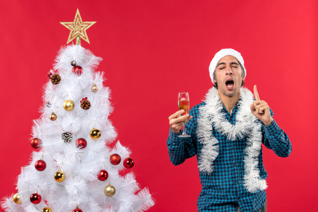 玻璃圣诞节心情自信坚定情绪化的年轻人戴着圣诞老人帽 穿着蓝色条纹衬衫 在圣诞树旁举起一杯葡萄酒人新年圣诞
