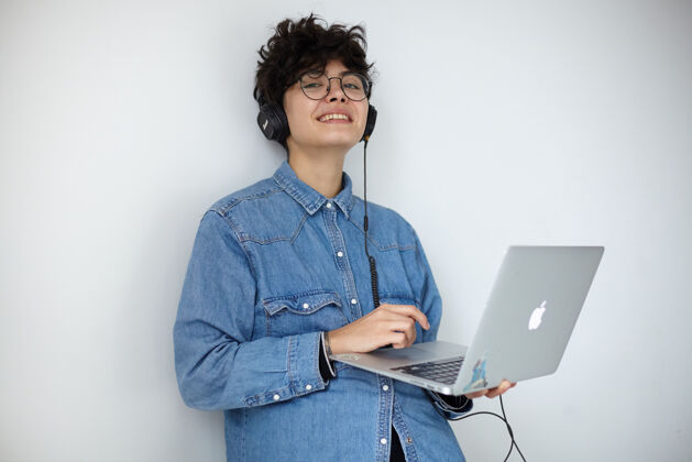 眼镜高兴的年轻黑发卷曲深褐色女士在蓝色牛仔裤衬衫听音频文件与耳机和微微微笑的相机 保持手提电脑 而站在白色的背景发型20多岁笔记本电脑