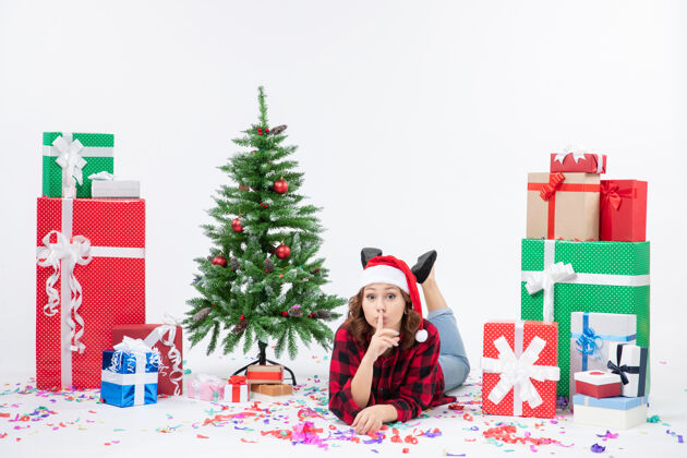 雪年轻女子躺在白色墙壁上的圣诞礼物和小圣诞树周围的正视图十二月人快乐