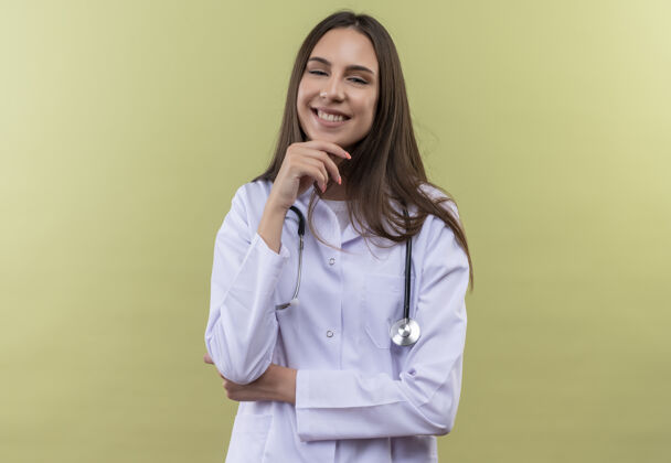 微笑微笑的年轻医生女孩穿着听诊器医用长袍 手放在绿色背景的下巴上下巴戴长袍
