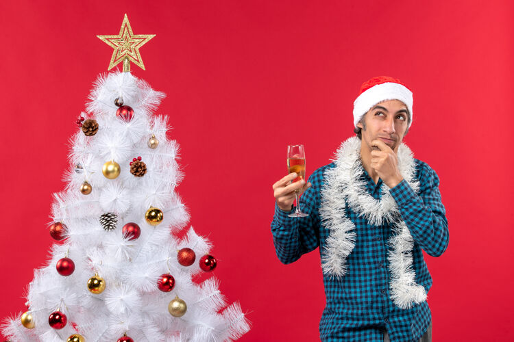 年轻人圣诞节心情快乐疯狂情绪混乱的年轻人戴着圣诞老人的帽子在一个蓝色条纹衬衫举起一杯葡萄酒靠近圣诞树新年情感装饰