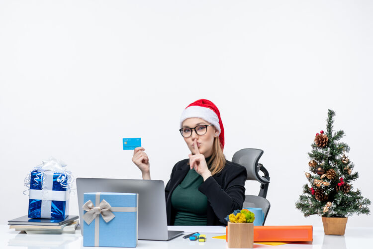 眼镜自信迷人的女人 戴着圣诞老人的帽子 戴着眼镜 坐在桌子旁 手里拿着银行卡 在办公室里做着沉默的手势圣诞节迷人笔记本电脑