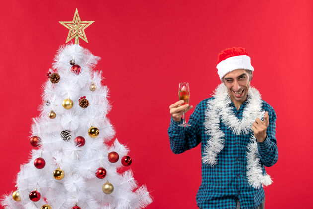疯狂圣诞心情快乐疯狂情绪好奇的年轻人戴着圣诞老人帽 穿着蓝色条纹衬衫 在圣诞树旁举起一杯葡萄酒年圣诞老人帽子