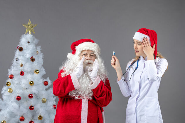 医生圣诞老人和女医生的前视图 女医生拿着测温仪在灰色的墙上快乐温度女性