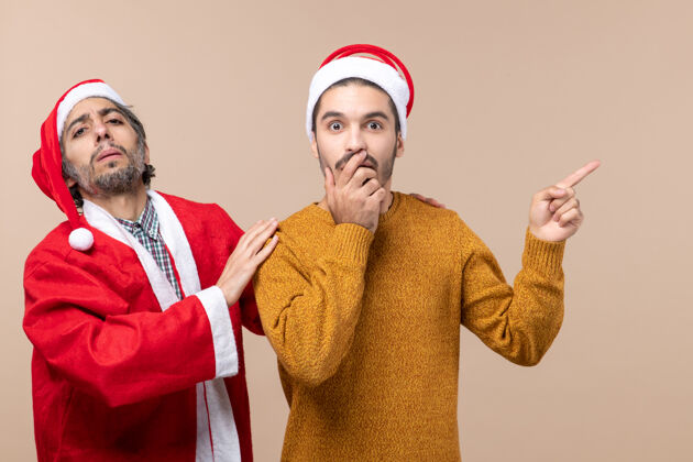 两个朋友正面图两个戴着圣诞帽的朋友一个抱着其他人的肩膀 另一个在米色孤立背景上显示方向手持显示帽子