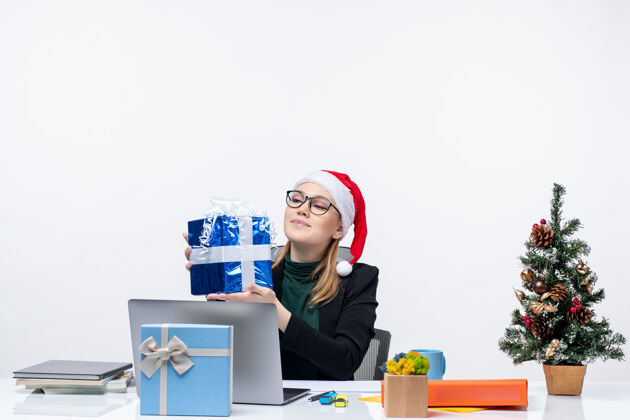 电脑圣诞气氛与戴着圣诞老人帽子和眼镜的年轻女子坐在一张桌子旁自豪地看着她的礼物白色背景圣诞节坐着圣诞老人