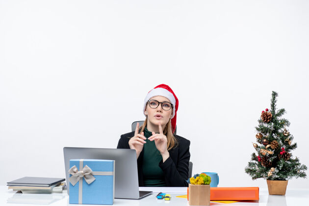 桌子紧张的女商人戴着圣诞老人的帽子坐在一张桌子旁 桌子上放着圣诞树和礼物 背景是白色的礼物秘书帽子