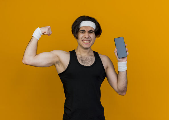 展示年轻的运动型男子穿着运动服和头带显示智能手机提高拳头显示二头肌看起来紧张头带拳头橙色