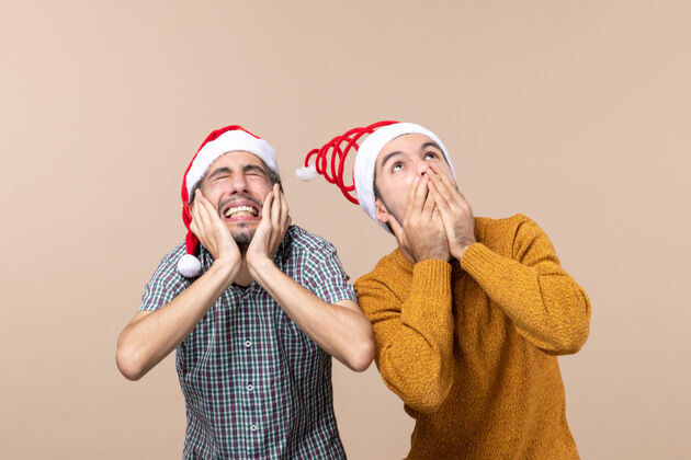 两个正面图两个戴着圣诞帽的吓坏了的家伙 一个捂着耳朵 另一个嘴巴在米色的孤立背景上帽子米色圣诞老人