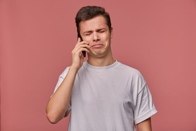 封闭一张摄影棚照片 一个心烦意乱的黑发男人穿着休闲服站在粉色背景下 手里拿着智能手机 脸上带着悲伤 在电话里听到坏消息发型通话男人