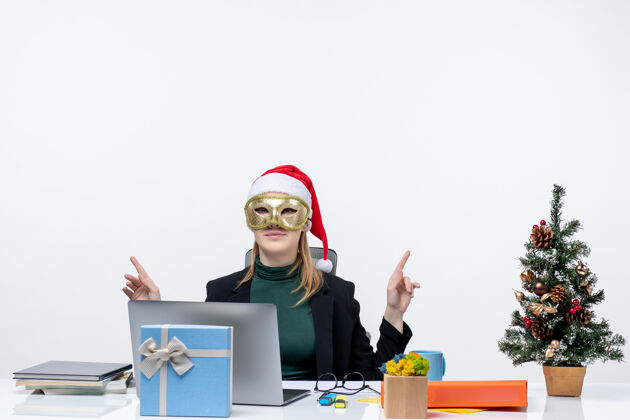 面具圣诞气氛 戴着圣诞老人帽 戴着面具的年轻女子坐在桌子旁 指着白色背景上的东西坐着笔记本电脑年轻