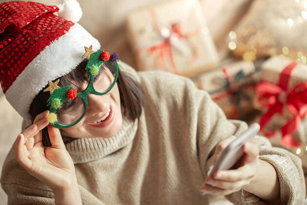 节日戴着滑稽的圣诞眼镜 戴着圣诞老人帽子 手里拿着电话的快乐女孩舒适有趣女人