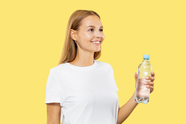 财务黄色工作室背景上的白人年轻女子半身像穿着白衬衫的漂亮女模特人类情感的概念 面部表情从瓶子里喝水成人人女性