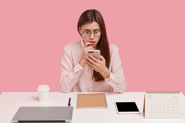 个人惊呆了的欧洲女人的照片在现代手机上浏览互联网 有惊恐的表情 在工作区摆姿势 穿着正式的衬衫工作区咖啡震惊