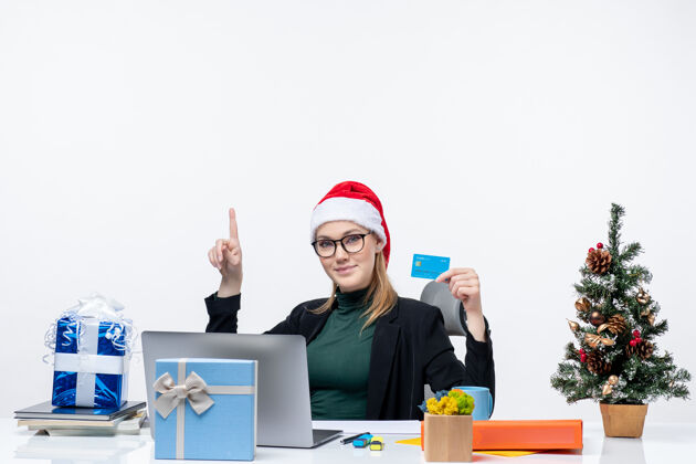 办公室积极迷人的女人戴着圣诞老人的帽子 戴着眼镜坐在桌旁 拿着圣诞礼物 指着办公室上方的银行卡迷人银行帽子