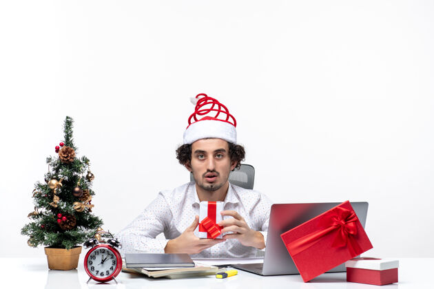 坐着圣诞气氛与年轻的商人与圣诞老人的帽子坐在办公室里 并举行了他的礼物摆出白色背景的相机圣诞老人年轻商人成人
