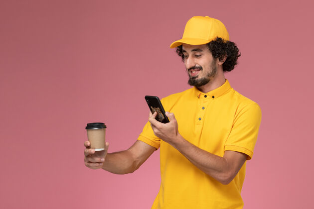 帽子正面图身穿黄色制服的男性信使和披风拿着送货咖啡杯在粉色墙上拍照咖啡男士快递