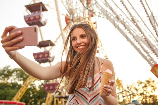 太阳镜快乐的年轻可爱的女士的肖像 带着迷人的微笑 在游乐园的景点上摆姿势 用智能手机拍照 手里拿着冰激凌筒温暖可爱冰