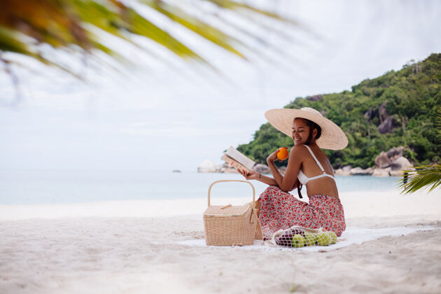 户外一个年轻女子戴着草帽 穿着白色针织衣服坐在沙滩地毯上天堂女人人