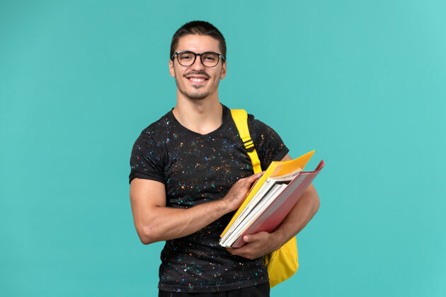 持有身穿深色t恤黄色背包的男学生正面图 浅蓝色墙上有文件和书籍男性人学生