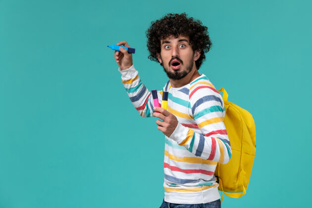 人穿着条纹衬衫 背着黄色背包 拿着毛毡笔的男生在蓝色墙上的正视图男性持有大学