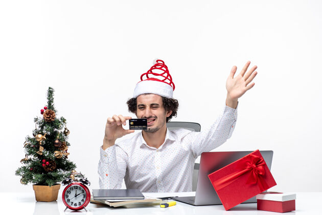 办公室节日喜庆的心情带着年轻疲惫愤怒的商人戴着圣诞老人的帽子 在白色背景的办公室里看着他的银行卡商务人士年轻商务