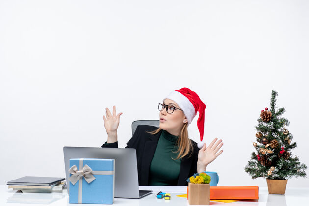电脑震惊的女商人戴着圣诞老人的帽子坐在一张桌子旁 桌子上有一棵圣诞树和一份礼物 上面是白色的背景帽子人笔记本电脑