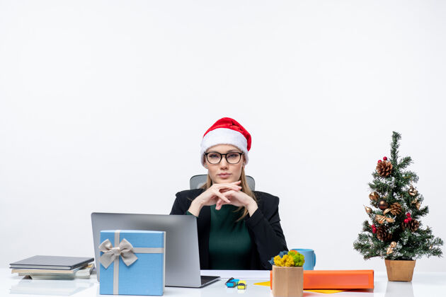 女士在白色背景的办公室里 一位自信的商务女士戴着圣诞老人帽坐在一张桌子旁 桌子上放着圣诞树和礼物礼物电脑办公室