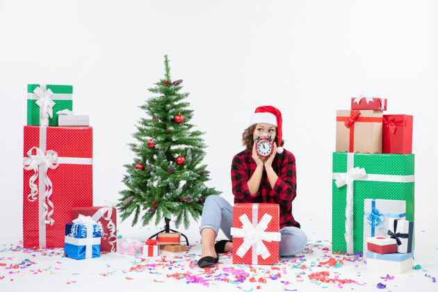 冬青前视图的年轻女子坐在圣诞礼物周围拿着白色墙上的时钟庆祝圣诞节玩具
