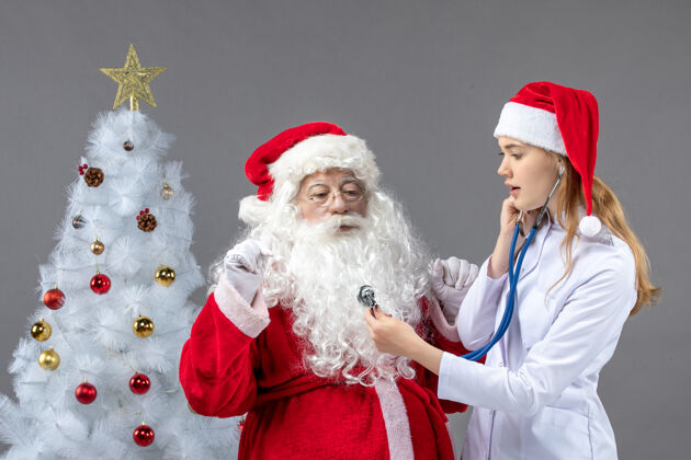 十二月圣诞老人和女医生的前视图 女医生正在灰墙上检查他的健康状况庆祝快乐圣诞老人