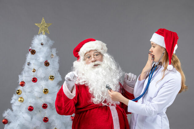 节日圣诞老人和女医生的前视图 女医生正在灰墙上检查他的健康状况庆祝圣诞老人快乐