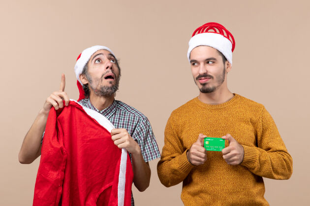 帽子前视图两个家伙一个拿着圣诞老人的外套 看着高处 另一个拿着信用卡 看着他的朋友在米色孤立的背景肖像男信用卡